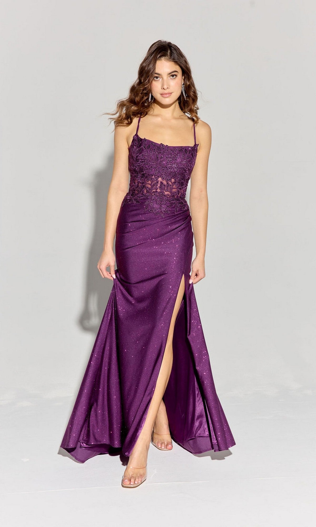 A woman models the Eureka EK129 long prom dress featuring a sleeveless lace bodice, floor-length glitter jersey sheath skirt with thigh-high slit, and lace-up open back, against a plain light background.